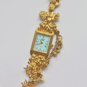 May include: Gold-toned quartz watch with a rectangular face and Roman numeral hour markers. The watch band features ornate cherub and star designs with rhinestone accents. A unique and decorative timepiece.