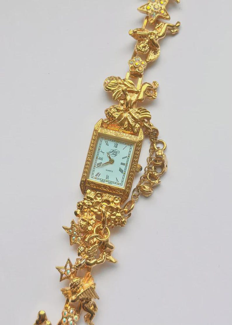 May include: Gold-toned quartz watch with a rectangular face and Roman numeral hour markers. The watch band features ornate cherub and star designs with rhinestone accents. A unique and decorative timepiece.