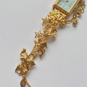 May include: A gold-toned quartz watch with a rectangular face and Roman numeral hour markers. The watch band features ornate cherub and star details with clear crystal accents. A delicate chain hangs from the band.