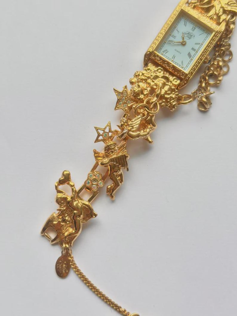 May include: A gold-toned quartz watch with a rectangular face and Roman numeral hour markers. The watch band features ornate cherub and star details with clear crystal accents. A delicate chain hangs from the band.