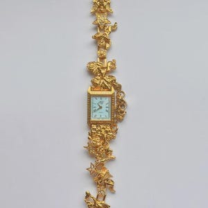 May include: A gold-toned wristwatch with a rectangular face and ornate detailing. The watch band features decorative figures and embellishments. The watch face has a light blue background with black Roman numerals and hands.