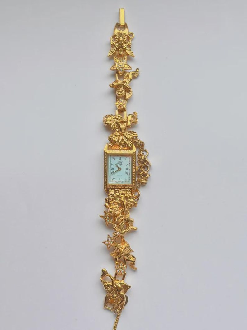 May include: A gold-toned wristwatch with a rectangular face and ornate detailing. The watch band features decorative figures and embellishments. The watch face has a light blue background with black Roman numerals and hands.