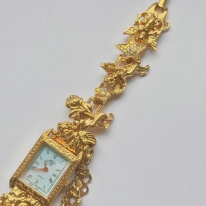 May include: Gold-toned quartz watch with a rectangular face and Roman numeral hour markers. The watch features ornate detailing, including cherubs, stars, and floral accents. The watch band is a series of gold chains.