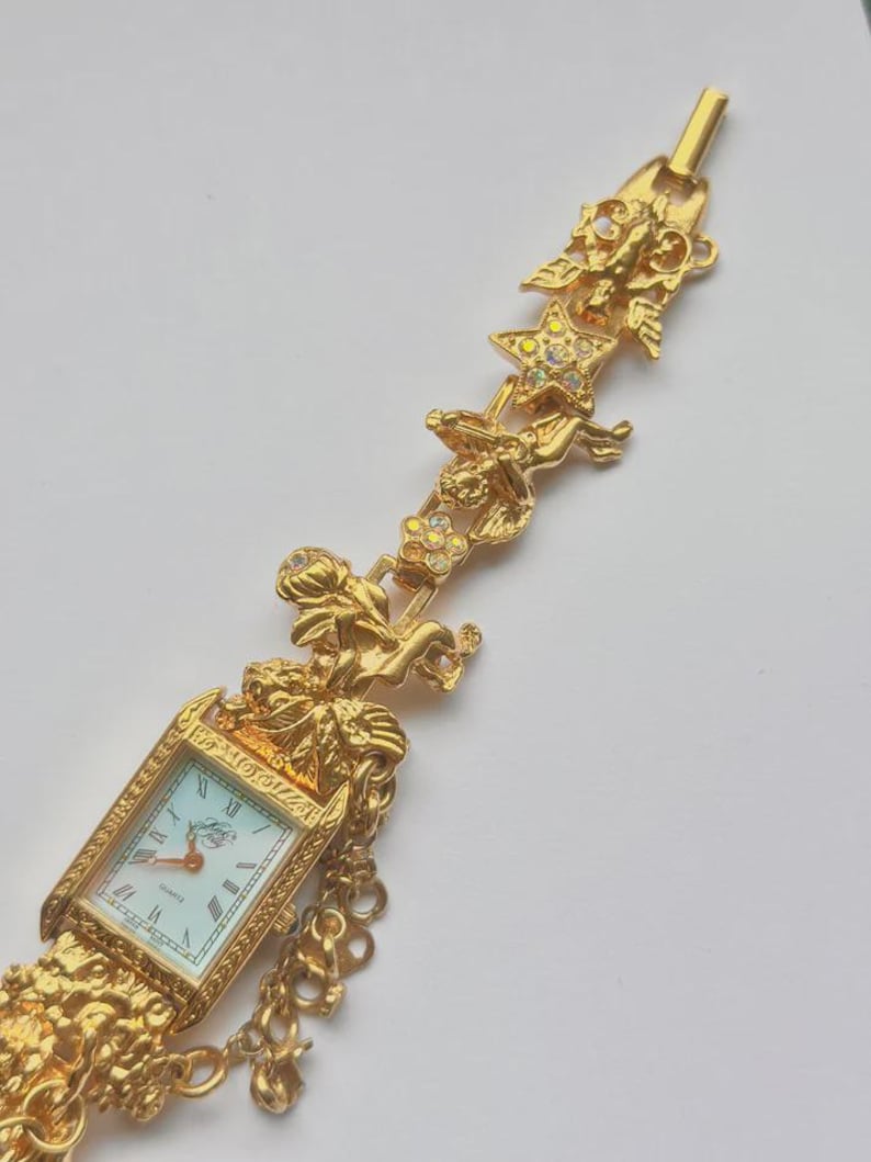 May include: Gold-toned quartz watch with a rectangular face and Roman numeral hour markers. The watch features ornate detailing, including cherubs, stars, and floral accents. The watch band is a series of gold chains.