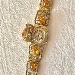 May include: A vintage gold-toned watch bracelet with amber-colored stones. The watch face is square and opens to reveal the time. The bracelet features filigree detailing and rectangular amber stones. The watch is displayed on a white, textured surface.