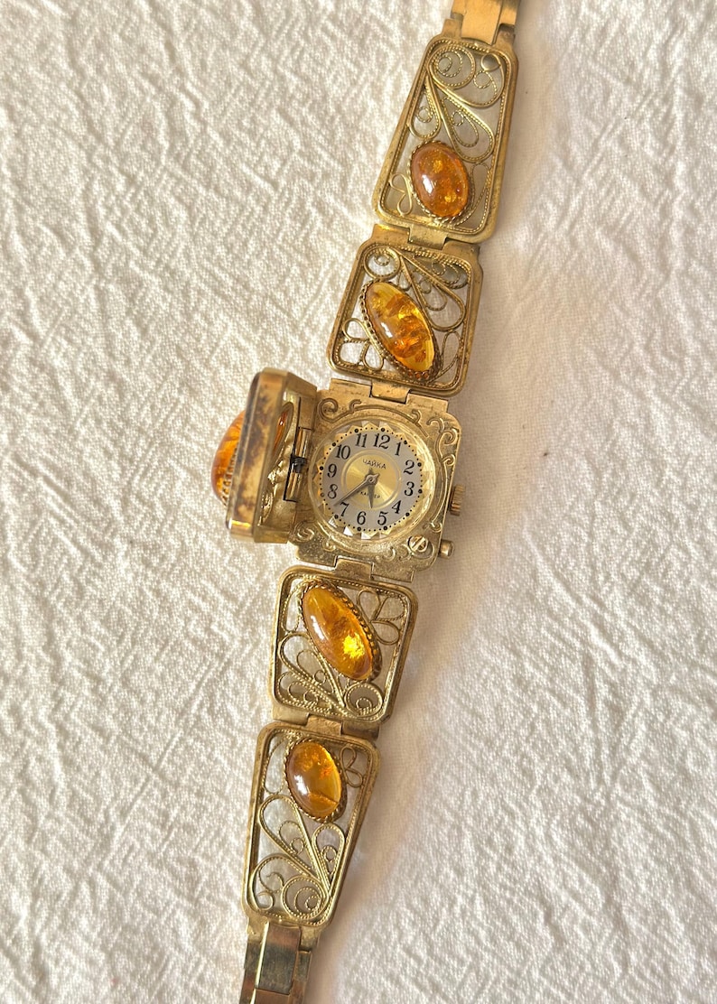 May include: A vintage gold-toned watch bracelet with amber-colored stones. The watch face is square and opens to reveal the time. The bracelet features filigree detailing and rectangular amber stones. The watch is displayed on a white, textured surface.