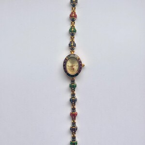 May include: Gold-toned wristwatch with an oval face and a decorative band. The watch face is gold with black hands and hour markers. The band features alternating red, green, and blue jewel-like accents. The watch is displayed against a white background.