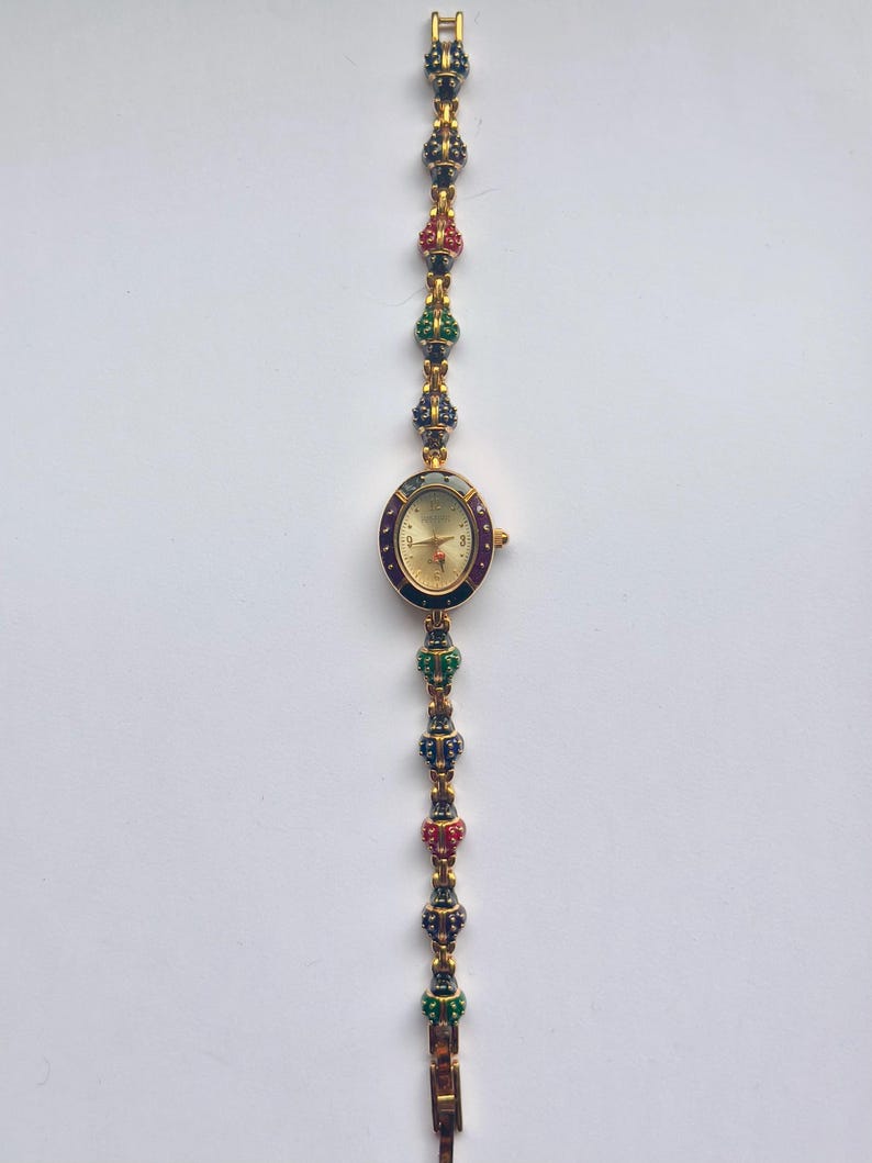 May include: Gold-toned wristwatch with an oval face and a decorative band. The watch face is gold with black hands and hour markers. The band features alternating red, green, and blue jewel-like accents. The watch is displayed against a white background.