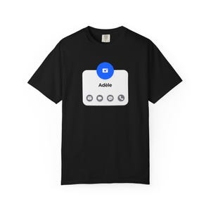 May include: Black t-shirt featuring a white graphic resembling a phone message. The graphic includes a blue circle with a camera icon, the name "Adèle", and several small icons below. The shirt is made of a soft material.
