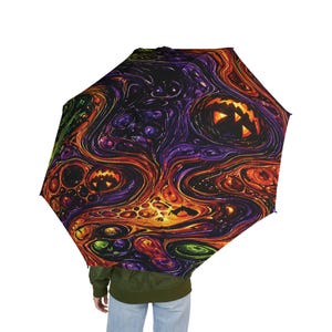 Peut inclure: Un parapluie ouvert avec un motif abstrait tourbillonnant dans les tons orange, violet, vert et noir, avec des visages de jack-o'-lantern. Le design est sur le thème d'Halloween. Le parapluie est ouvert, révélant le motif.