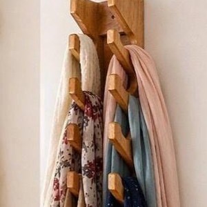 May include: A vertical wooden wall-mounted rack with multiple pegs holding various scarves. The rack features a light brown wood grain finish and is organized with a cream scarf, a floral print scarf, a pink scarf, and a blue scarf.