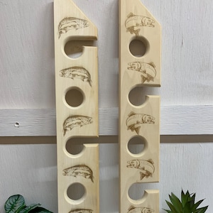 May include: Two vertical light-colored wooden fishing rod racks featuring laser-engraved illustrations of trout. Each rack contains four circular cutouts for rod storage and notched side slots for securing fishing equipment in place.