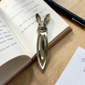 May include: A gold-coloured metal bookmark with a rabbit head design and red gem eyes. The bookmark is placed in an open book. A black pen lies on the wooden surface next to the book.