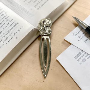 May include: A silver-toned metal bookmark featuring a sculpted dog head at the top and a long, pointed, open-centre blade design. The bookmark is positioned vertically on a surface next to an open book with printed text and a silver ballpoint pen.