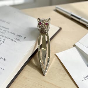 Può includere: Un segnalibro in metallo color argento con una testa di gatto scolpita sulla parte superiore, dotata di due piccole pietre rotonde rosa acceso come occhi. Il corpo del segnalibro è lungo, sottile e appuntito con un'apertura centrale.