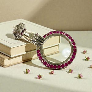 May include: A silver-toned magnifying glass with a hand-shaped handle and a circular lens. The rim of the lens is adorned with small, round, red gemstones. The handle rests on a stack of books, with small pink flowers scattered around.