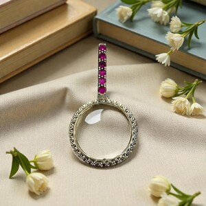 May include: A silver magnifying glass with a clear lens and a handle set with red gemstones. The round frame has an ornate, detailed design. The magnifying glass is on a beige surface, with books and flowers in the background.