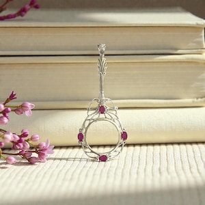 May include: A silver-toned pendant with a decorative design, featuring a central open circle and six oval-shaped red gemstones. The pendant has an ornate handle and is displayed on a textured surface with a backdrop of books and pink flowers.