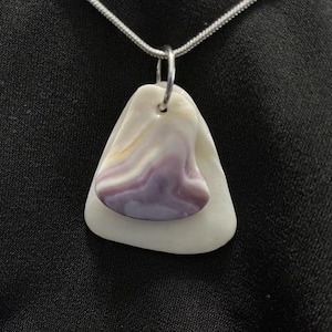 May include: A pendant necklace featuring a white and purple seashell pendant. The pendant is made of two pieces, a larger white piece and a smaller purple piece. The necklace has a silver chain. The background is a dark fabric.