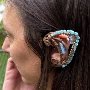 May include: A brown ear cuff with a woven design, adorned with light blue beads and a central blue gemstone. The cuff is worn on the ear, creating an elven or fantasy aesthetic.