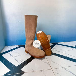 Puede incluir: Una bota alta de ante beige con tacón plano y punta redonda. La suela es de cuero marrón y cuenta con una placa metálica en la punta con tachuelas visibles. Una etiqueta redonda blanca con el texto Caligar Craftico está en la caña de la bota.