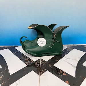 May include: A pair of green leather elf shoes with a curled toe and pointed flaps. The shoes have a lace-up closure and are set against a blue and white background. The text "Caligar Craftco" is on the shoe.