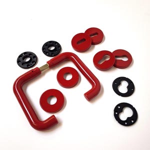 Puede incluir: Un conjunto de componentes de herrajes para puertas rojos y negros, que incluyen manijas, cubiertas de cerraduras y placas de montaje. El hardware está dispuesto sobre una superficie blanca, mostrando un diseño moderno. Medidas en cm.