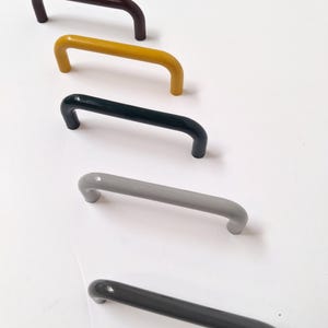 May include: Five cabinet pulls in a row, each with a curved handle design. The colors are, from top to bottom, dark brown, yellow, dark green, gray, and dark gray. The pulls are made of a solid material and have a smooth, glossy finish.