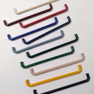 Może przedstawiać: Zbiór dziesięciu metalowych uchwytów w kształcie litery U w różnych kolorach, w tym kremowym, bordowym, czerwonym, niebieskim, granatowym, szarobłękitnym, zielonym, beżowym, żółtym i czarnym. Uchwyty są równomiernie rozmieszczone i ułożone po przekątnej.