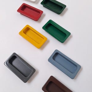 Puede incluir: Una variedad de tiradores empotrados de armario de colores, en tonos como blanco, rojo, amarillo, verde, gris, azul y marrón. Los tiradores rectangulares tienen un interior curvado, diseñado para un agarre fácil.