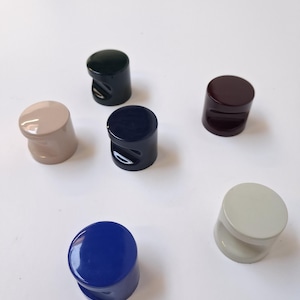 Könnte beinhalten: Sechs farbige, zylindrische Flaschenverschlüsse in verschiedenen Farbtönen, darunter Blau, Grün und Beige. Jeder Verschluss hat eine abgerundete Oberseite und einen gebogenen Griff. Die Verschlüsse sind auf einer weißen Oberfläche angeordnet.