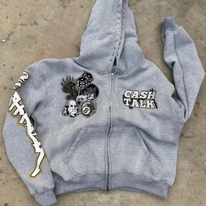 Puede incluir: Una sudadera con capucha gris con cremallera y una capucha adornada con pedrería. La sudadera con capucha presenta parches con gráficos temáticos de dinero y el texto "CASH TALK". La manga izquierda tiene texto en un diseño dorado y blanco.