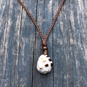 May include: A handcrafted necklace featuring a white seashell pendant with brown spots, suspended from a braided brown cord. The pendant is attached to the cord with a small wooden bead. The necklace is displayed against a weathered, blue-gray wooden surface.