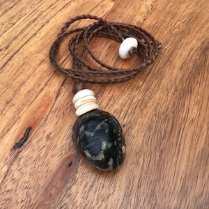 May include: A handcrafted necklace featuring a dark, patterned pendant with a braided brown cord. The necklace includes white beads and a dark, oval-shaped pendant. The necklace rests on a wooden surface.