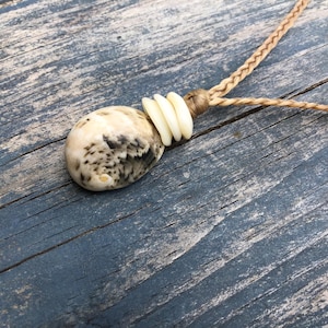 May include: A necklace featuring a smooth, mottled beige and brown stone pendant accented by three small, off-white circular beads. The pendant is attached to a braided tan cord that extends upward, showcasing a simple and functional design.