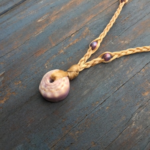 May include: A necklace featuring a single round, purple-toned seashell pendant attached to a braided tan cord. The cord includes two small, dark purple spherical beads integrated into the braid, extending upward from the central knot.