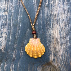 May include: A seashell pendant necklace with a braided brown cord. The pendant is a yellow and orange scallop shell. A small wooden bead accents the cord. The necklace rests on a blue wooden surface.
