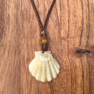 May include: A seashell pendant necklace with a braided brown cord. The pendant is a cream-colored scallop shell. A wooden bead is strung on the cord above the shell. The necklace is displayed on a wooden surface.