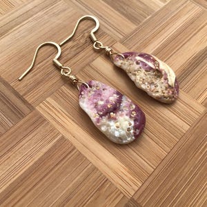 May include: A pair of drop earrings featuring natural, irregular shell fragments with a mottled purple and white color palette. Each shell is attached to a gold-toned metal hook by a small wire-wrapped loop, showcasing a textured, porous surface.
