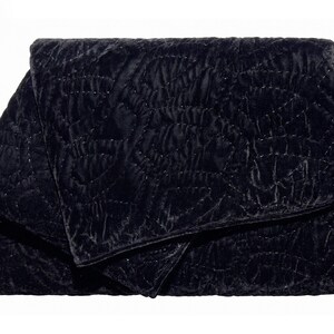 Puede incluir: Un clutch de terciopelo negro plegado con un patr&oacute;n acolchado. La tela tiene un aspecto suave y texturizado, y las costuras crean un dise&ntilde;o geom&eacute;trico sutil. El clutch es rectangular con una solapa plegada.