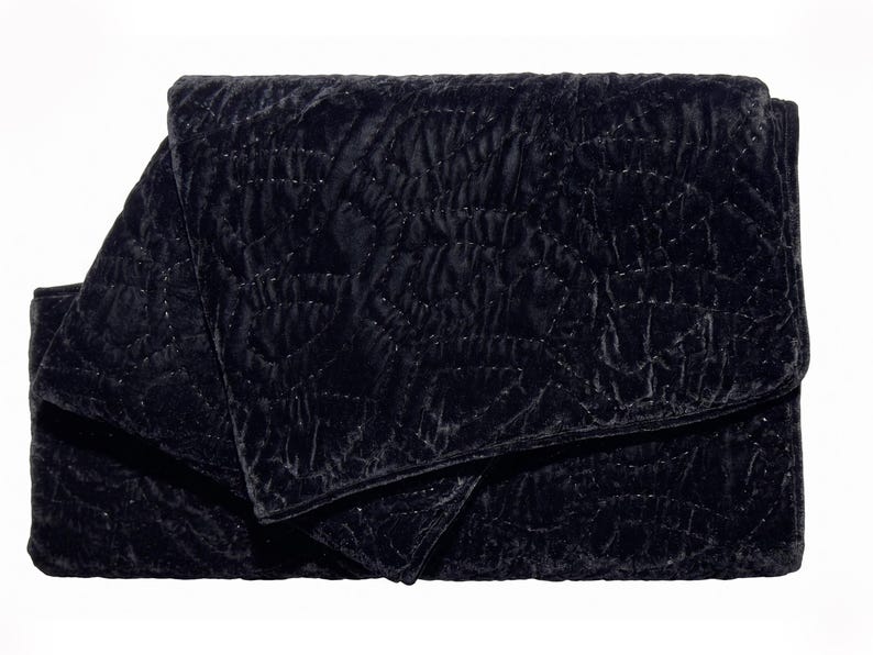 Puede incluir: Un clutch de terciopelo negro plegado con un patr&oacute;n acolchado. La tela tiene un aspecto suave y texturizado, y las costuras crean un dise&ntilde;o geom&eacute;trico sutil. El clutch es rectangular con una solapa plegada.