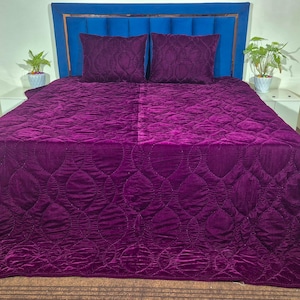 Peut inclure: Un couvre-lit en velours violet profond avec un motif circulaire matelassé, accompagné de deux oreillers carrés assortis. La literie est placée contre une tête de lit bleue rembourrée avec des coutures verticales et des bordures métalliques.