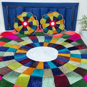 May include: A bedspread featuring a circular, multi-coloured patchwork design in velvet fabric with a large white centre circle. Two matching round, multi-coloured velvet pillows with white centres are placed against a dark blue upholstered headboard.