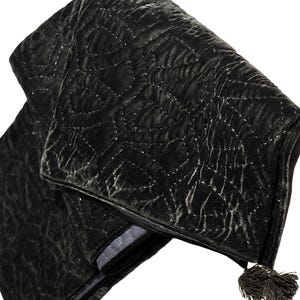 Puede incluir: Un clutch de terciopelo negro plegado con un patr&oacute;n acolchado y una borla. El clutch tiene un forro gris plateado y un dise&ntilde;o cosido decorativo. La est&eacute;tica general es elegante y sofisticada.