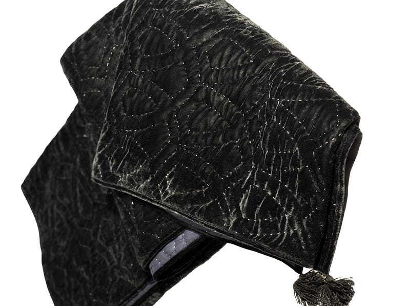 Puede incluir: Un clutch de terciopelo negro plegado con un patr&oacute;n acolchado y una borla. El clutch tiene un forro gris plateado y un dise&ntilde;o cosido decorativo. La est&eacute;tica general es elegante y sofisticada.