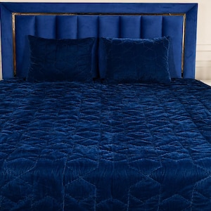 May include: A deep blue velvet bedspread and two matching rectangular pillows featuring a stitched geometric pattern. The bedding set is displayed on a bed with a tall, vertical-channeled blue velvet headboard with gold metal trim accents.