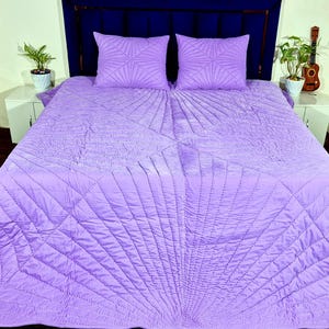 Op de afbeelding: Een lavendelkleurige sprei en twee bijpassende rechthoekige kussens met een gestikt waaiervormig patroon dat vanuit het midden van de quilt en de kussenslopen uitstraalt, wat zorgt voor een textuur op de gehele beddengoedset.