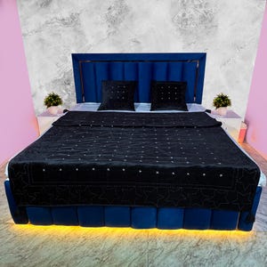 May include: A modern bed with a blue velvet headboard and base. The bed is made up with a black quilted blanket and two black square pillows. The bed frame has a built-in yellow light.