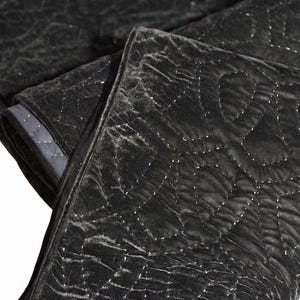 Puede incluir: Manta acolchada negra doblada con un patr&oacute;n de diamantes. La manta tiene una textura suave y aterciopelada y un borde gris oscuro. Las costuras son visibles, creando una superficie texturizada. La manta est&aacute; doblada sobre s&iacute; misma.