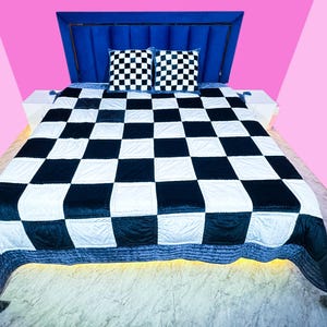 May include: A bed featuring a blue headboard and a black and white chequered quilt. Two chequered pillows are on the bed. The background is pink and white.